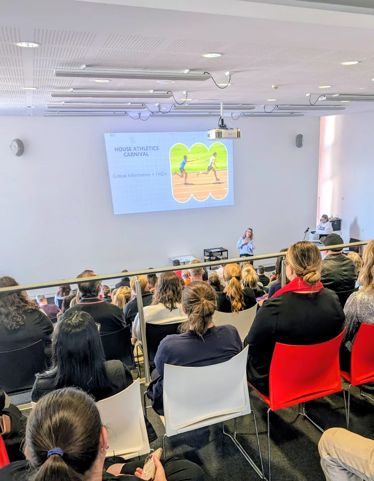 Audience watches a presentation about a house athletics carnival, relevant for understanding sports-related injuries and the importance of chiropractic care for athletes' performance and injury prevention.