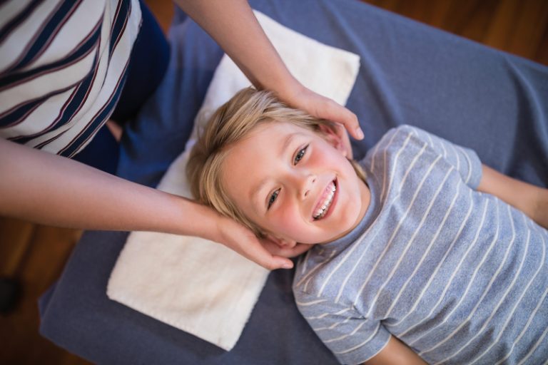 Smiling young boy receiving gentle chiropractic treatment on his neck and head, lying on a treatment table.