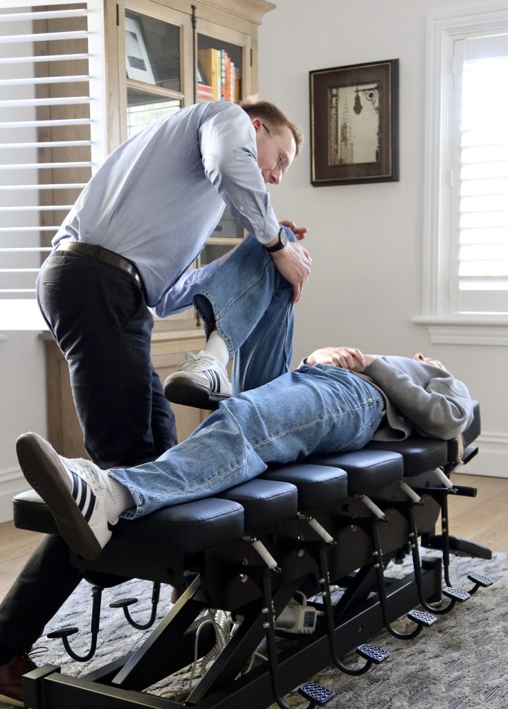 Chiropractor adjusting a patient's leg on a specialized chiropractic table in a well-lit office.
