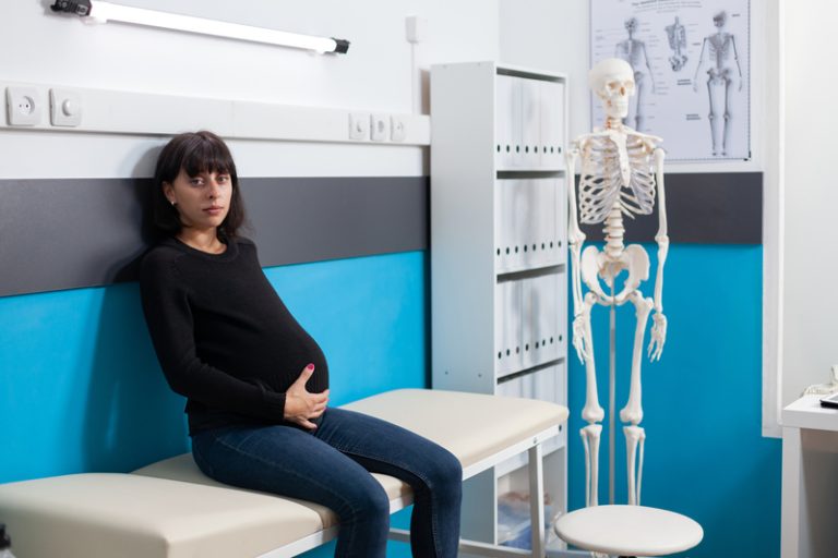 Pregnant woman awaits chiropractic treatment in a clinic, holding her belly with concern.