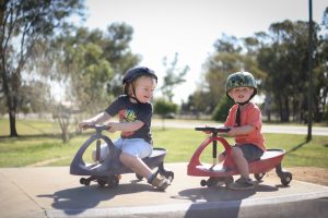 Two young children wearing helmets enjoy riding wiggle cars, promoting healthy spine movement and core strength, concepts often addressed in pediatric chiropractic care.